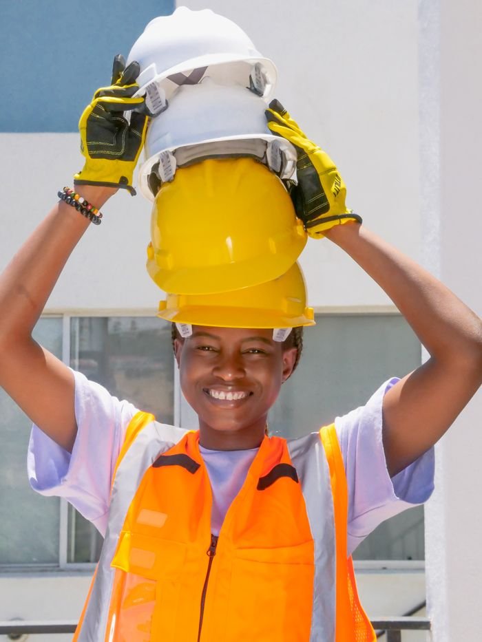 A cheerful woman engineer holding multiple safety helmets, wearing protective gear and smiling.